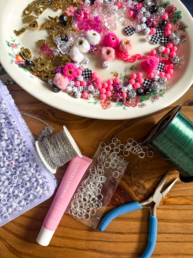 Supplies for DIY lip gloss charms featuring pink and checkered beads, gold trinkets, jewelry chain, and jump rings in a floral vintage bowl.