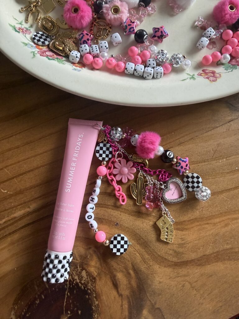 Supplies for DIY lip gloss charms featuring pink and checkered beads, gold trinkets, jewelry chain, and jump rings in a floral vintage bowl.