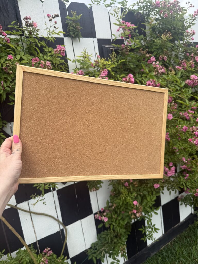 hand holding a rectangular corkboard in front of greenery and pink flowers, with a black-and-white checkered surface in the background.