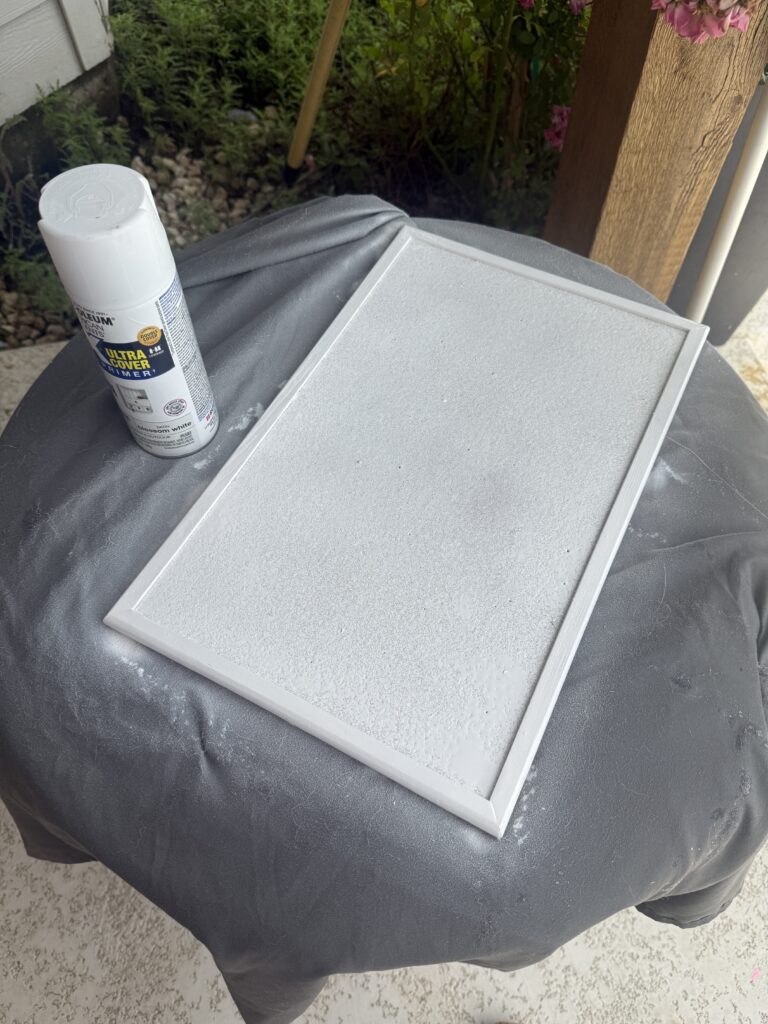 a corkboard frame freshly spray-painted white, resting on a covered surface next to a can of spray paint.