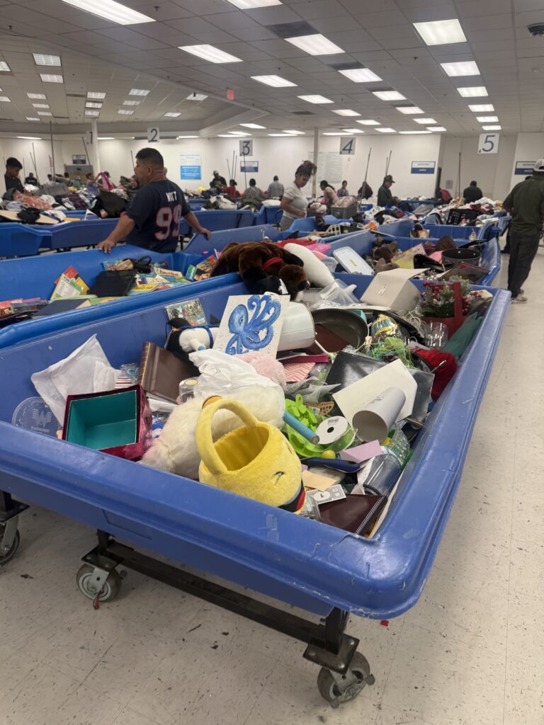 Crowded Houston Goodwill outlet bins filled with assorted clothing as shoppers search through large blue bins in a busy thrift store warehouse setting.