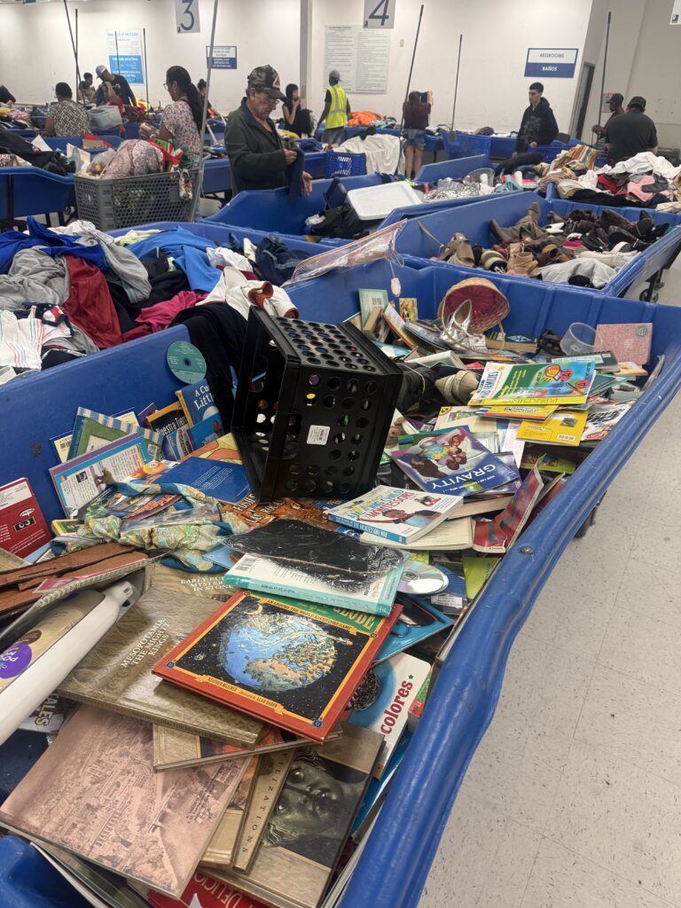 Crowded Houston Goodwill outlet bins filled with assorted clothing as shoppers search through large blue bins in a busy thrift store warehouse setting.