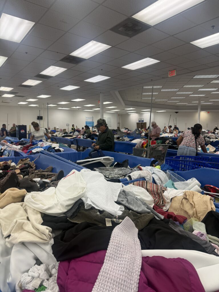 Crowded Houston Goodwill outlet bins filled with assorted clothing as shoppers search through large blue bins in a busy thrift store warehouse setting.