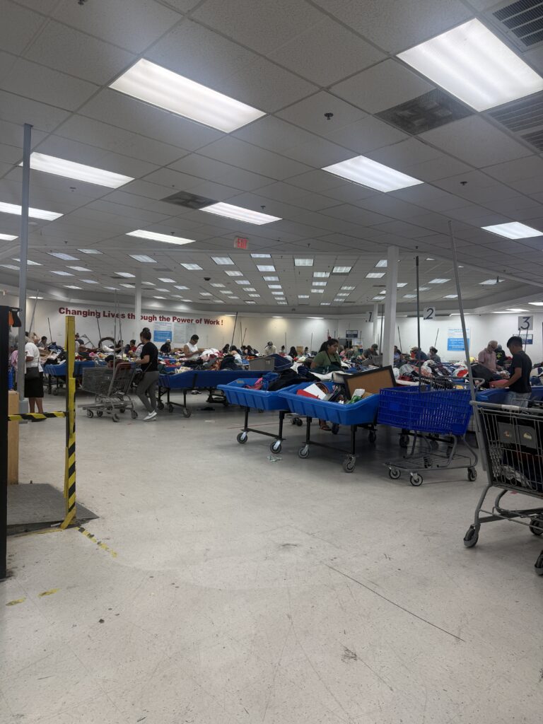 Crowded Houston Goodwill outlet bins filled with assorted clothing as shoppers search through large blue bins in a busy thrift store warehouse setting.