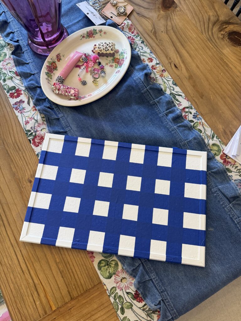 DIY setup: a white frame taped in a blue-and-white checkered pattern on a table, alongside a plate of craft supplies, a purple cup, and denim fabric.