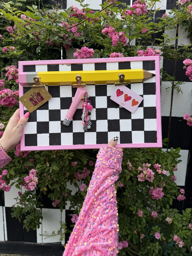 hand holding a pink-framed black-and-white checkered memo board featuring a yellow pencil holder and clipped notes, set against pink flowers and greenery.