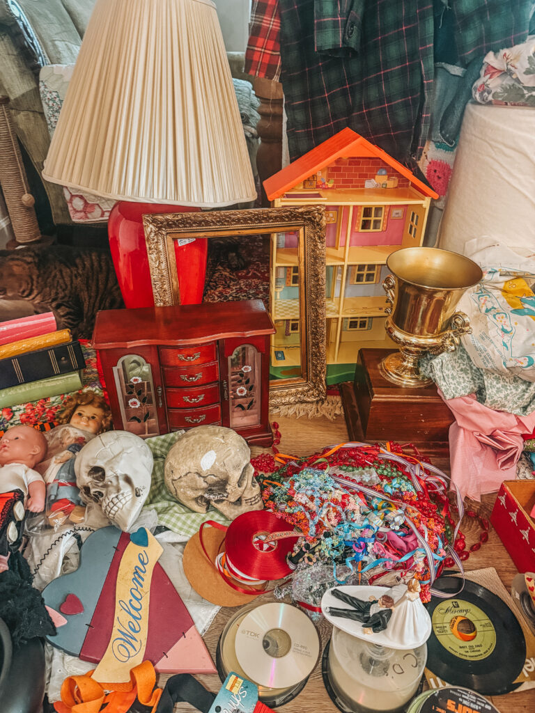 Detailed vignette of thrifted decor including a dollhouse, framed art, lamp, brass vase, and small skull accents arranged in a maximalist vintage style.