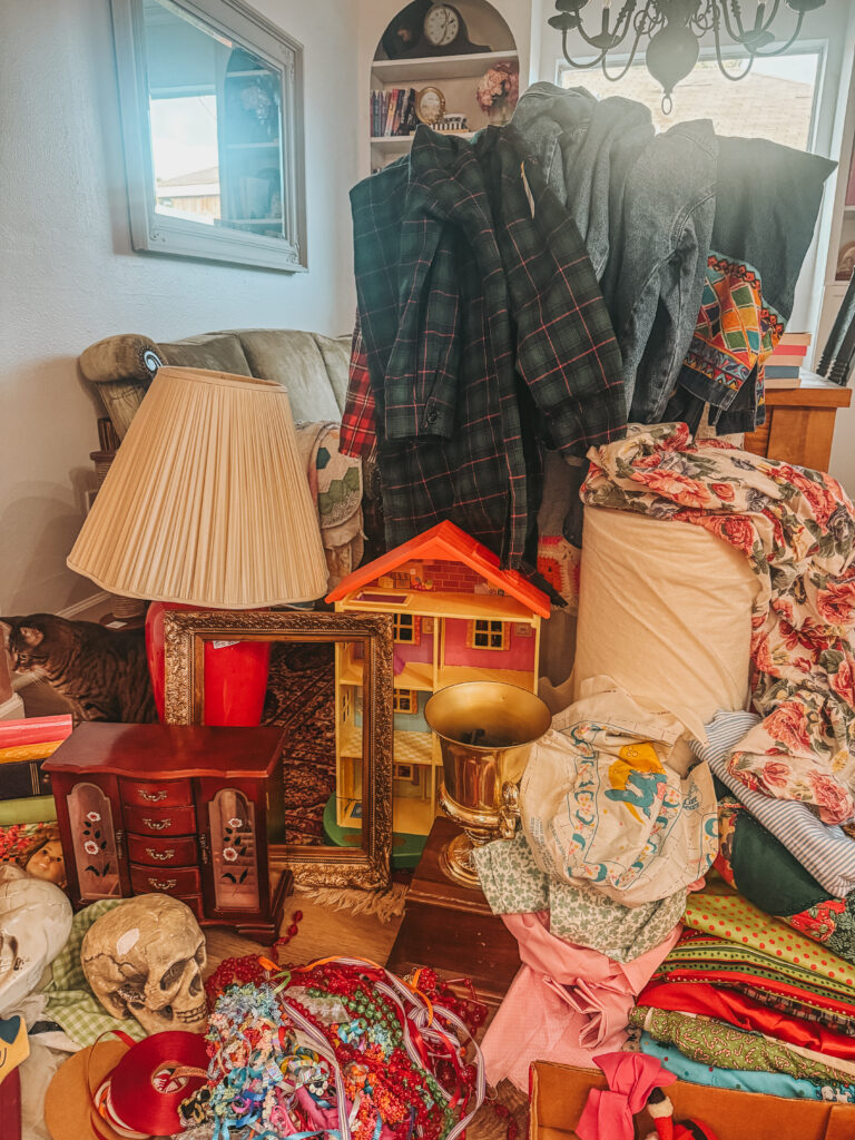 Cozy thrifted home decor setup with a table lamp, dollhouse, folded fabrics, and layered vintage textiles sourced from Houston Goodwill outlet bins.