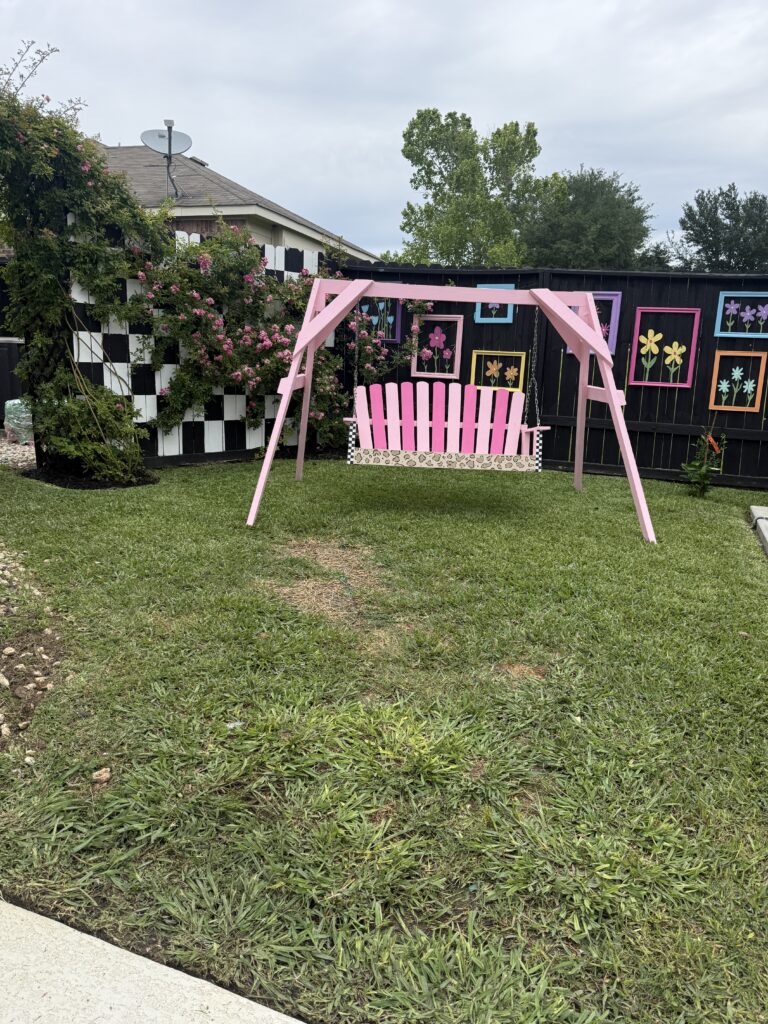 swing set painted a bold neon pink with a floral patterned seat, positioned in front of a fence decorated with bright, multicolored flower frames and a black-and-white checkered privacy screen.