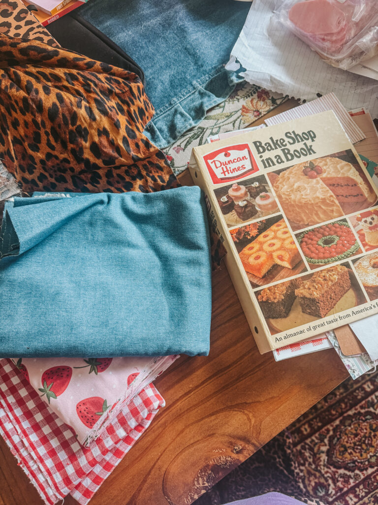 Crafting materials laid out for a DIY recipe junk journal project, including blue denim, leopard print fabric, and a vintage 'Duncan Hines Bake Shop in a Book' cookbook.