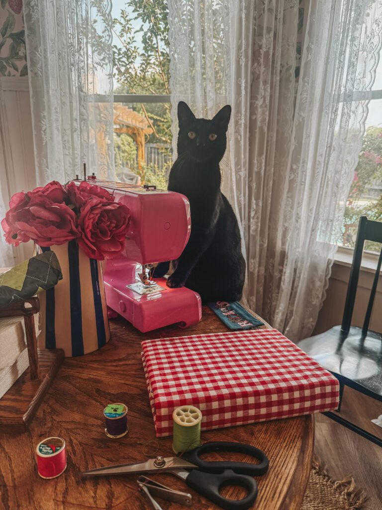 A black cat sitting on a craft table looking at the camera, situated behind a pink sewing machine and a denim pocket decorated with fabric letters spelling 'RECIPES'.