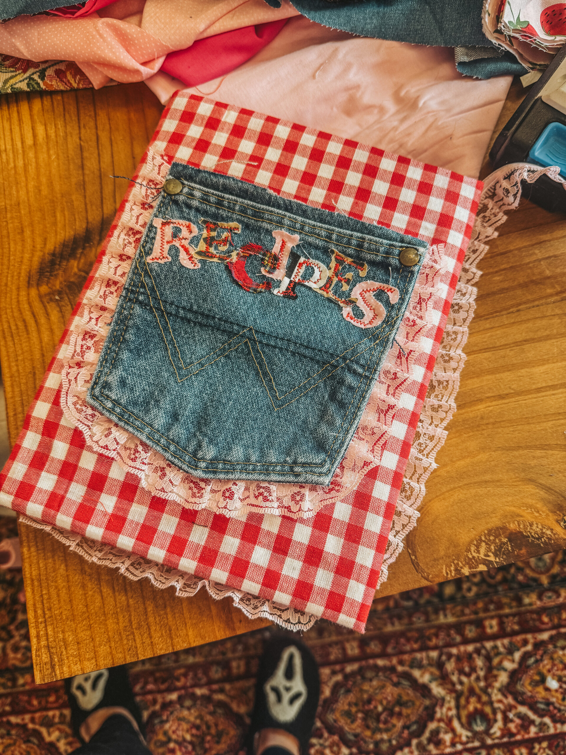 Front cover of a handmade recipe junk journal featuring red and white gingham fabric. A repurposed denim pocket is centered on the front, with the word 'RECIPES' spelled out in colorful, patterned fabric letters.