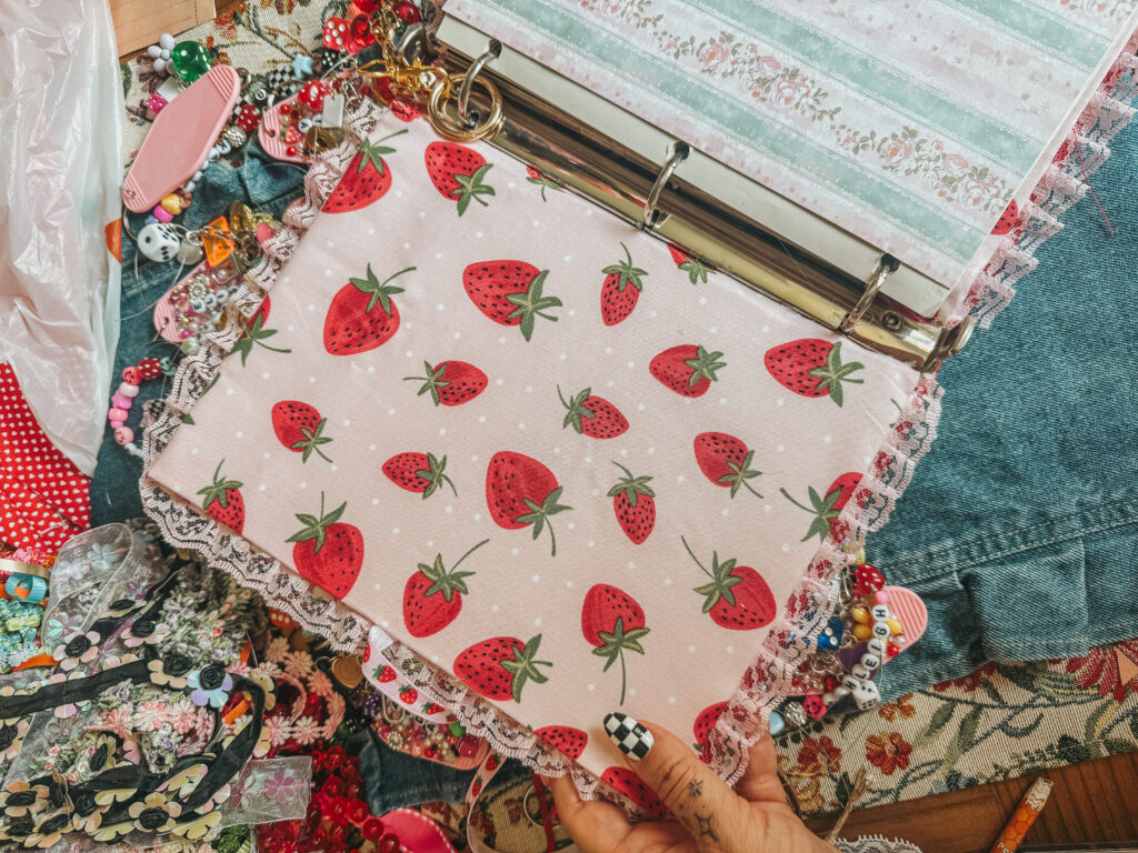 Inside cover of a DIY recipe junk journal featuring pink strawberry-patterned fabric, lace trim, decorative charms, and a silver three-ring binder mechanism.
