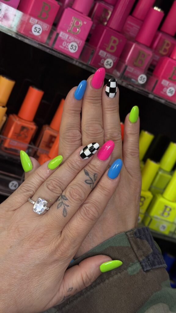 A vibrant, mismatched manicure featuring neon green, bright blue, hot pink, and black-and-white checkerboard almond-shaped nails, shown alongside a large diamond engagement ring and dainty finger tattoos.