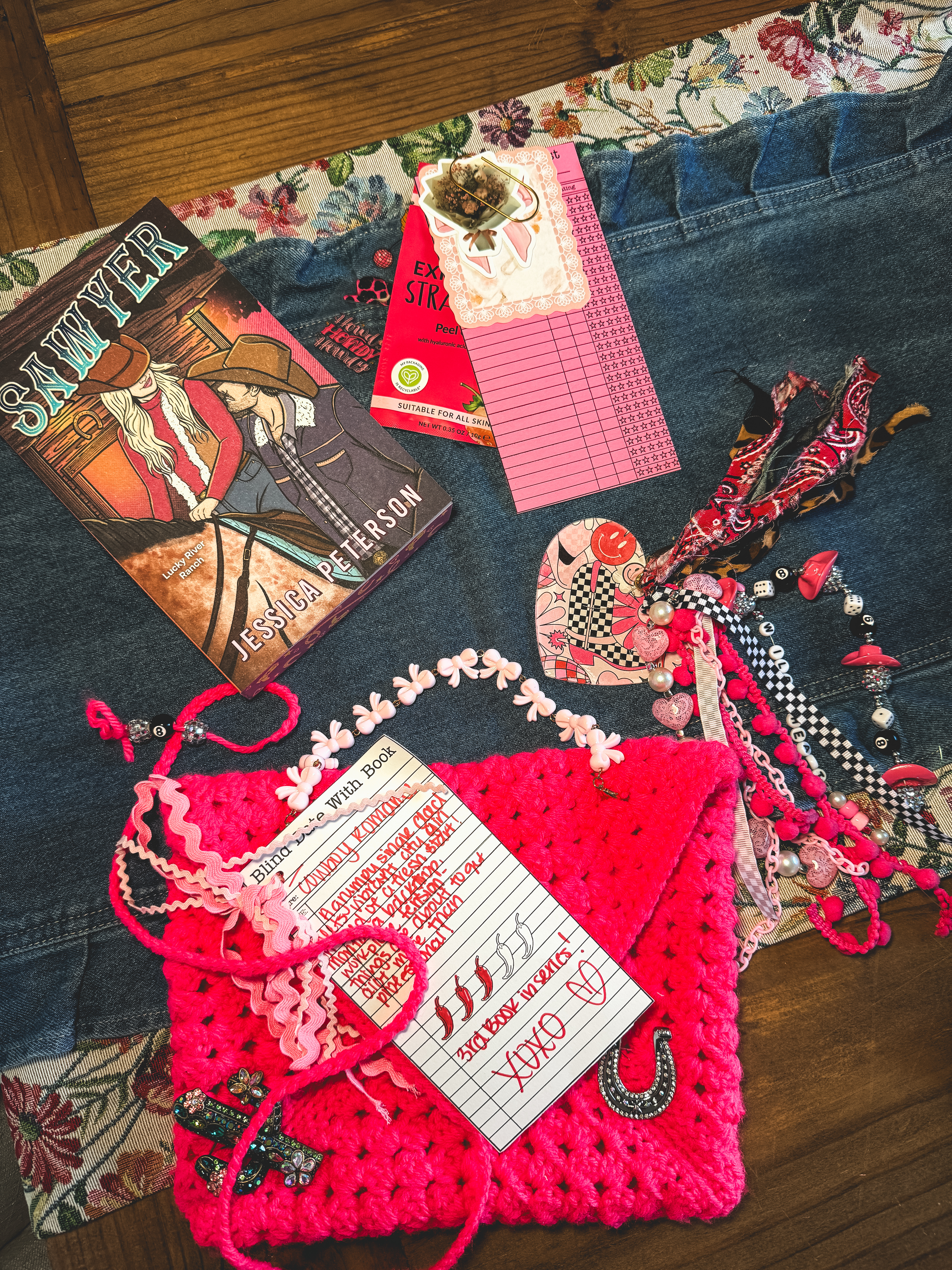 A bright pink crocheted pouch holding a handwritten "Blind Date with a Book" card for a country romance, placed next to the book "Sawyer" by Jessica Peterson and a pink library due-date card.