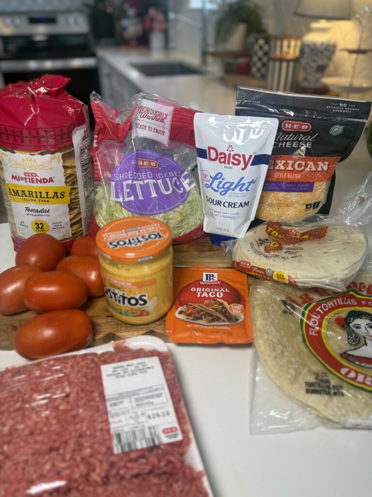 Groceries laid out on a counter for making Crunchwraps, including packages of ground beef, flour tortillas, yellow corn tostadas, shredded iceberg lettuce, Mexican style blend cheese, sour cream, a jar of salsa con queso, a packet of taco seasoning, and fresh tomatoes.