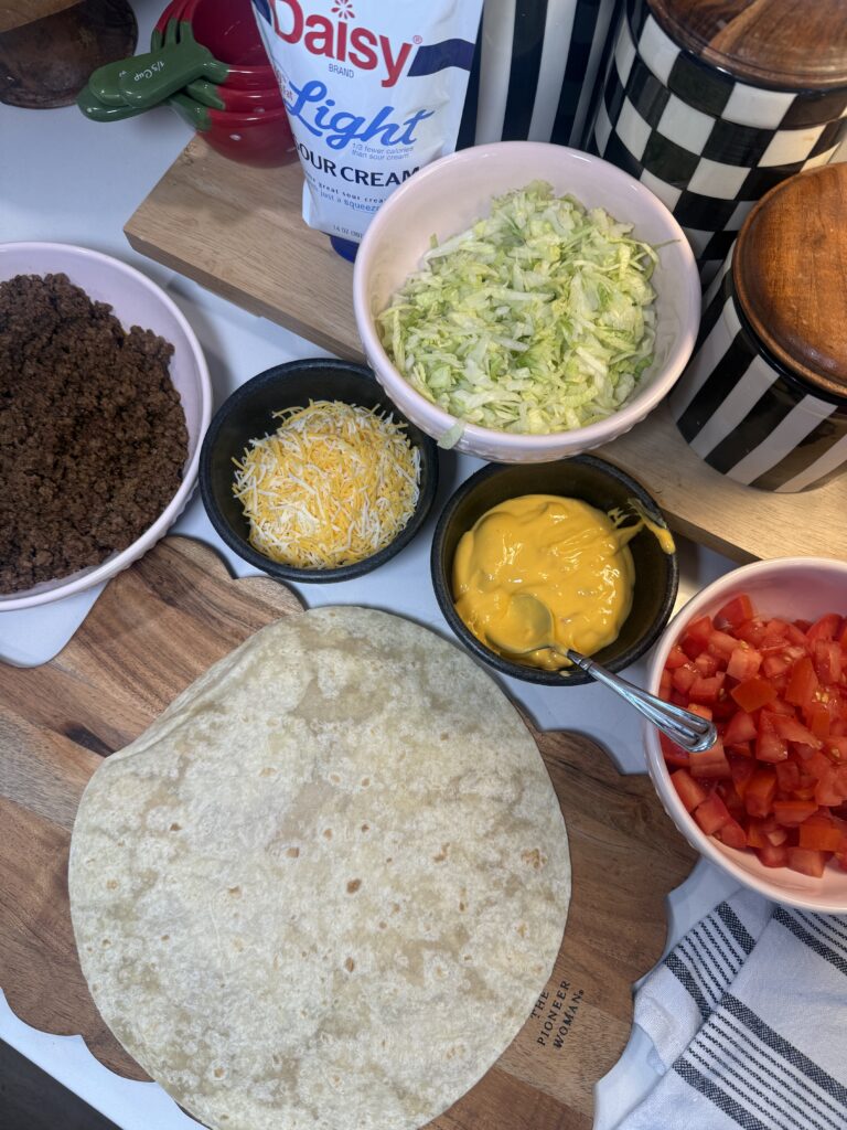 Top-down view of ingredients for a Crunchwrap on a wooden board, including a large flour tortilla, and individual bowls filled with seasoned ground beef, shredded cheese, nacho cheese sauce, shredded lettuce, and diced tomatoes.