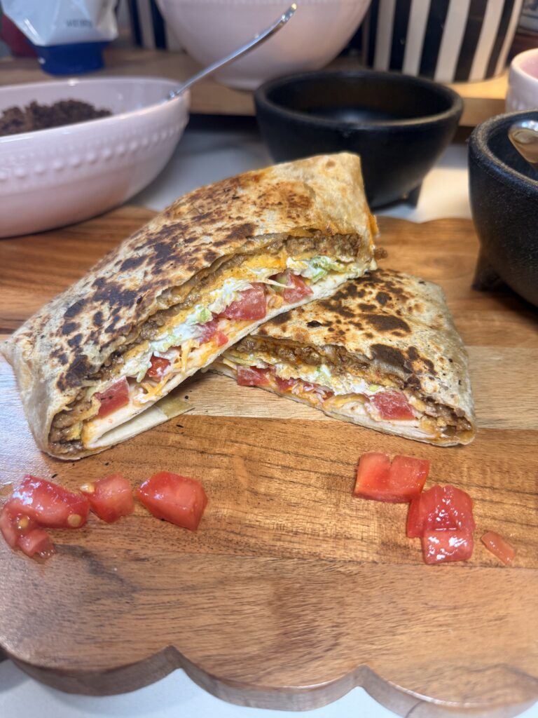 A halved, toasted Crunchwrap resting on a wooden cutting board next to a small white bowl. Scattered diced tomatoes.