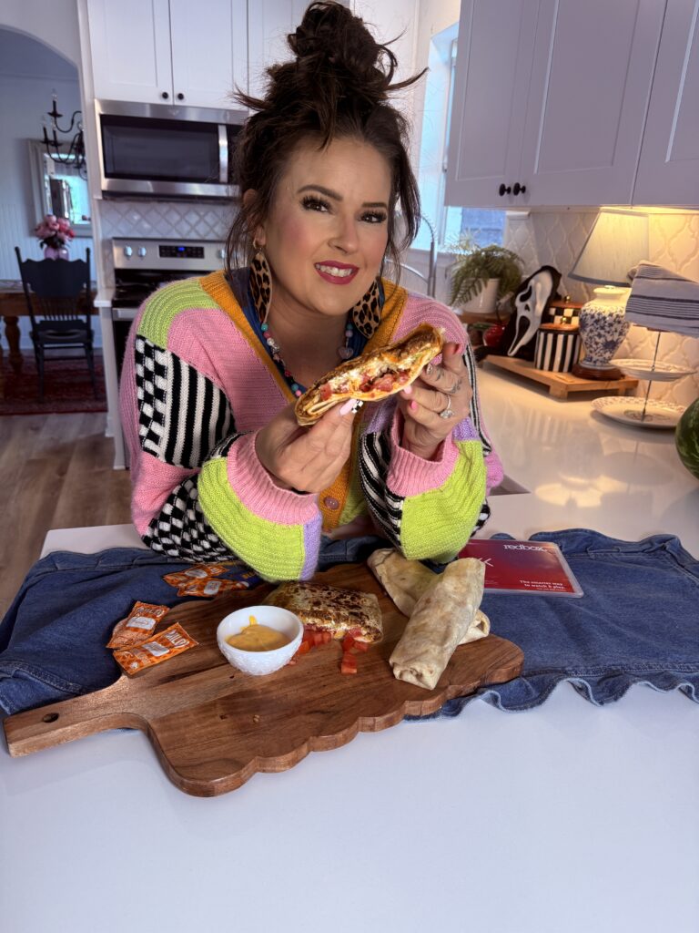 A smiling woman wearing a colorful color-block cardigan holds half of a Crunchwrap in her kitchen. In front of her is a wooden serving board with the other half of the Crunchwrap, burritos, cheese sauce, and hot sauce packets resting on a denim runner.