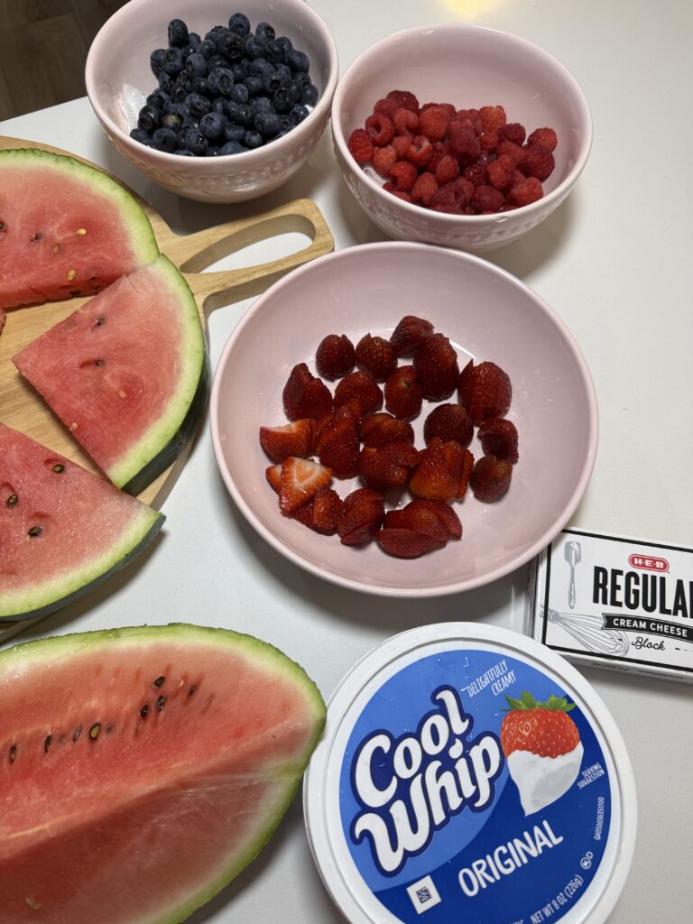 Ingredients for making watermelon fruit pizza, featuring fresh plain watermelon slices, a bowl of sliced strawberries, a tub of Cool Whip, and a block of cream cheese.