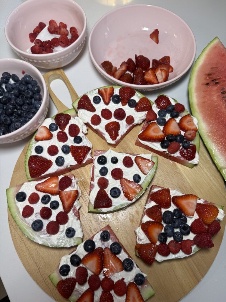 Close-up of watermelon slices on a wooden cutting board spread with white cream frosting and topped with fresh strawberries, blueberries, and raspberries to look like fruit pizza.