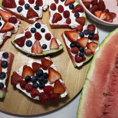 Close-up of watermelon slices on a wooden cutting board spread with white cream frosting and topped with fresh strawberries, blueberries, and raspberries to look like fruit pizza.