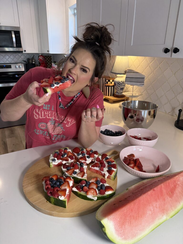 A woman standing in a kitchen taking a bite out of a slice of decorated watermelon fruit pizza, with the remaining slices and bowls of fresh berries on the counter in front of her.
