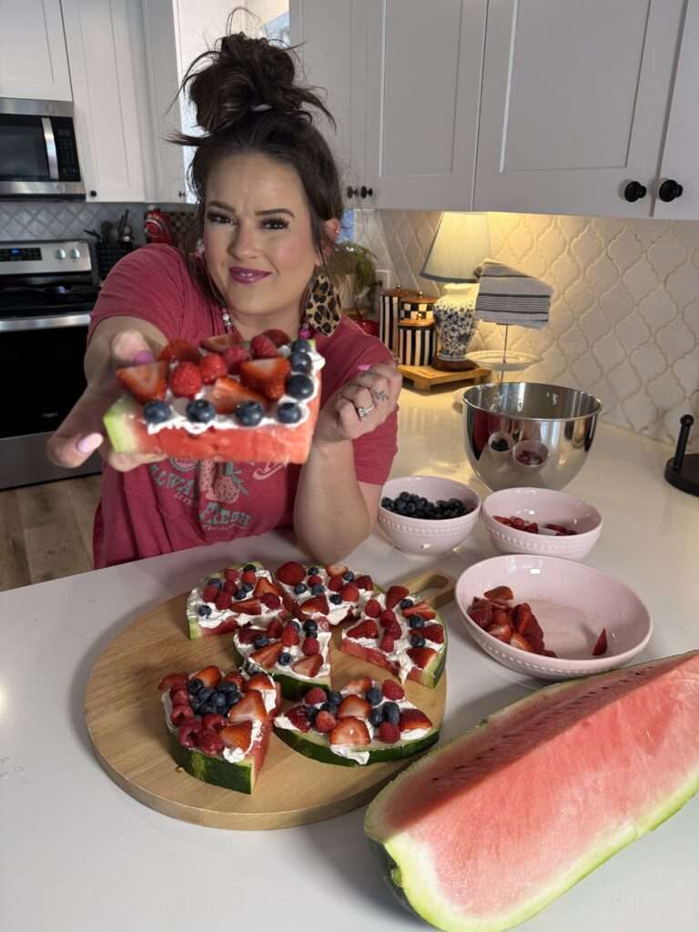 A woman standing in a kitchen taking a bite out of a slice of decorated watermelon fruit pizza, with the remaining slices and bowls of fresh berries on the counter in front of her.