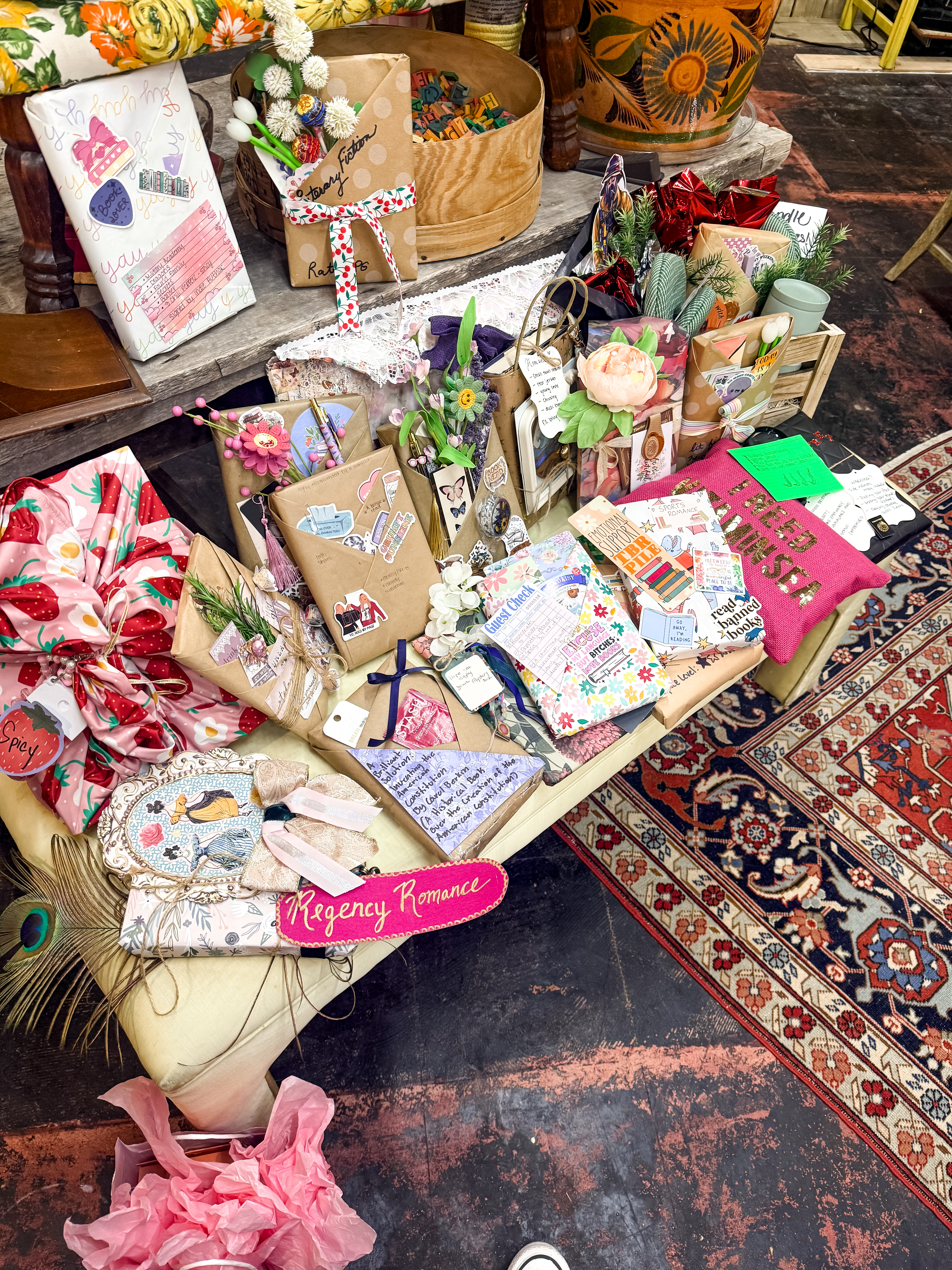 A wide shot of a highly decorated collection of "blind date with a book" gifts, beautifully wrapped in colorful paper, strawberry patterned fabric, and ribbons, scattered across a dark patterned rug.