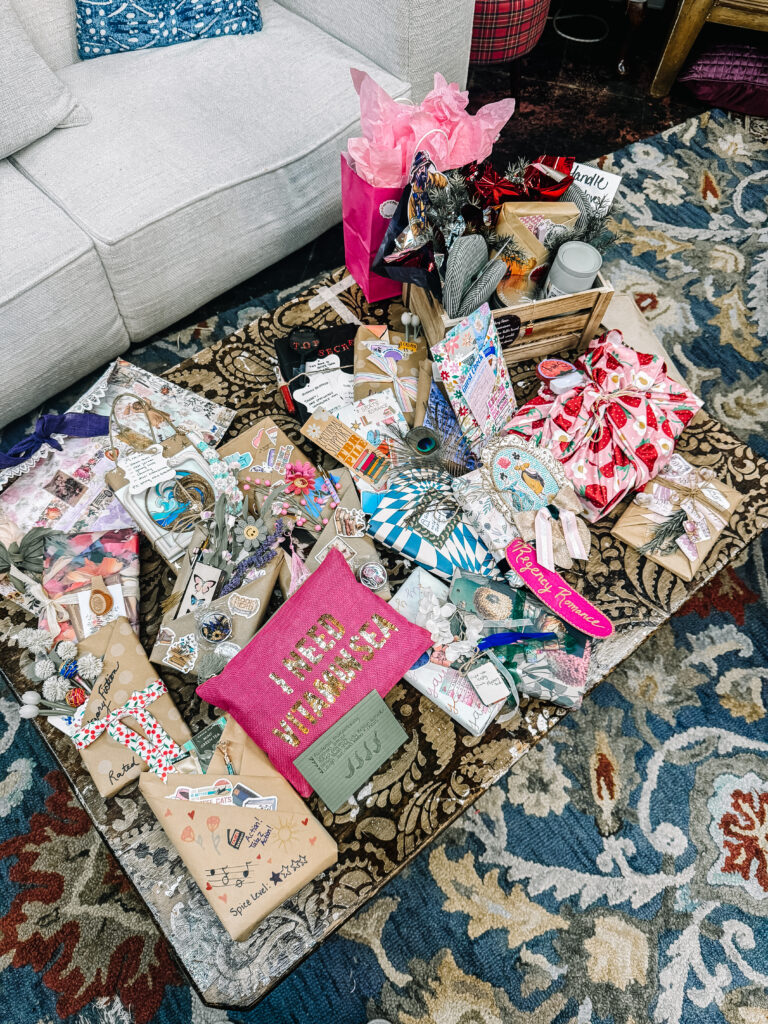 A maximalist display of intricately wrapped books for a summer-themed "blind date with a book," featuring various patterned wrapping papers, flowers, and a bright pink pouch that says "I NEED VITAMIN SEA."