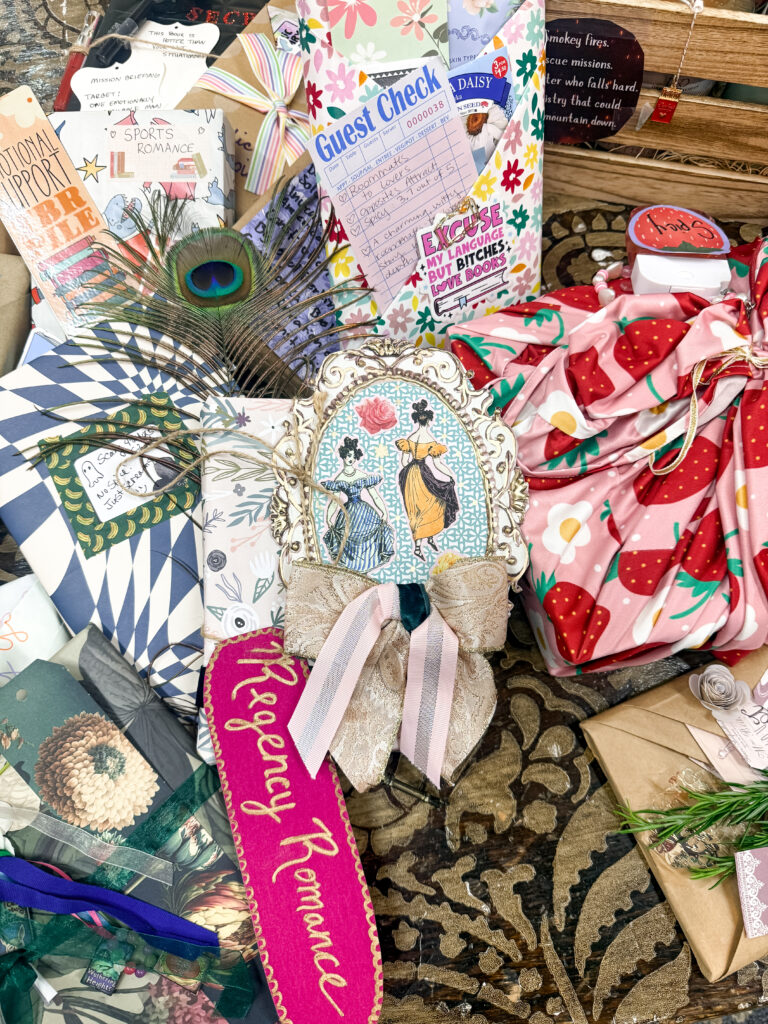 A close-up of beautifully wrapped books, including one adorned with a decorative cameo frame of women in historical dresses for a Regency Romance theme, next to a pink strawberry fabric wrap.