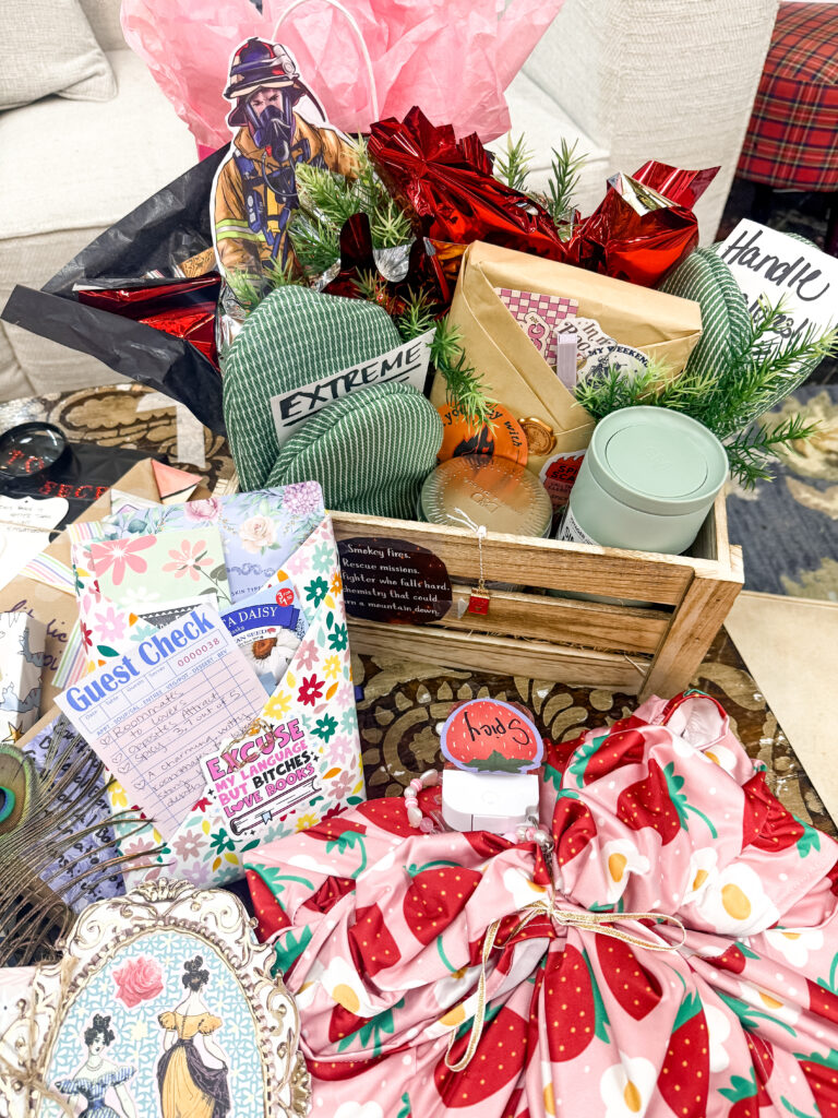 A firefighter-themed book gift basket in a small wooden crate, featuring a firefighter cutout, green pot holders, and a wrapped book inside a floral envelope with a book rating guest check.