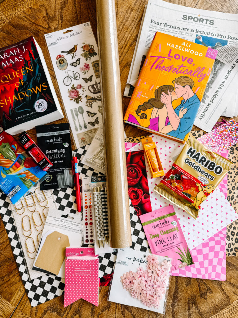 A flat lay of book lover gift items including the novels "Queen of Shadows" and "Love, Theoretically," alongside Haribo gummy bears, pink clay face masks, and decorative stationery on a wooden table.