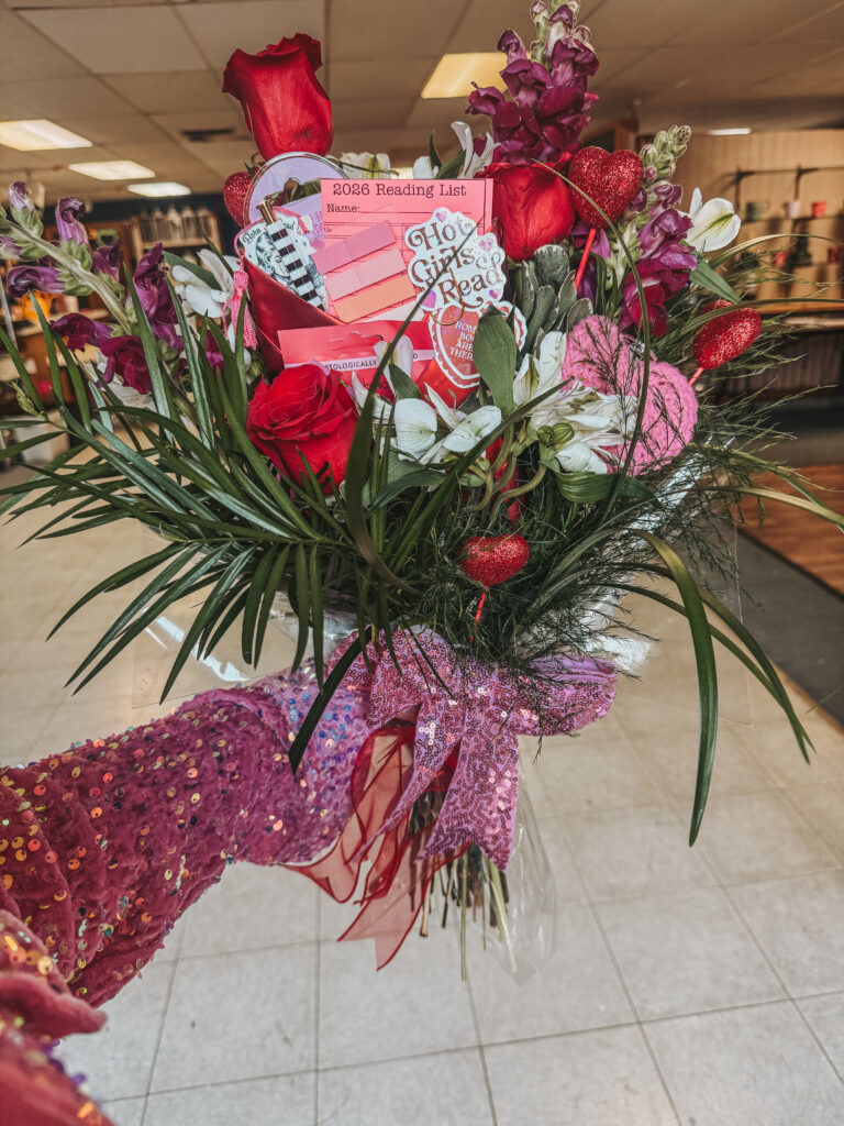 A person wearing a sparkly pink sequined sleeve holding a large, festive bouquet of red roses, with bookish gifts like a "2026 Reading List" card and "Hot Girls Read" stickers tucked inside.